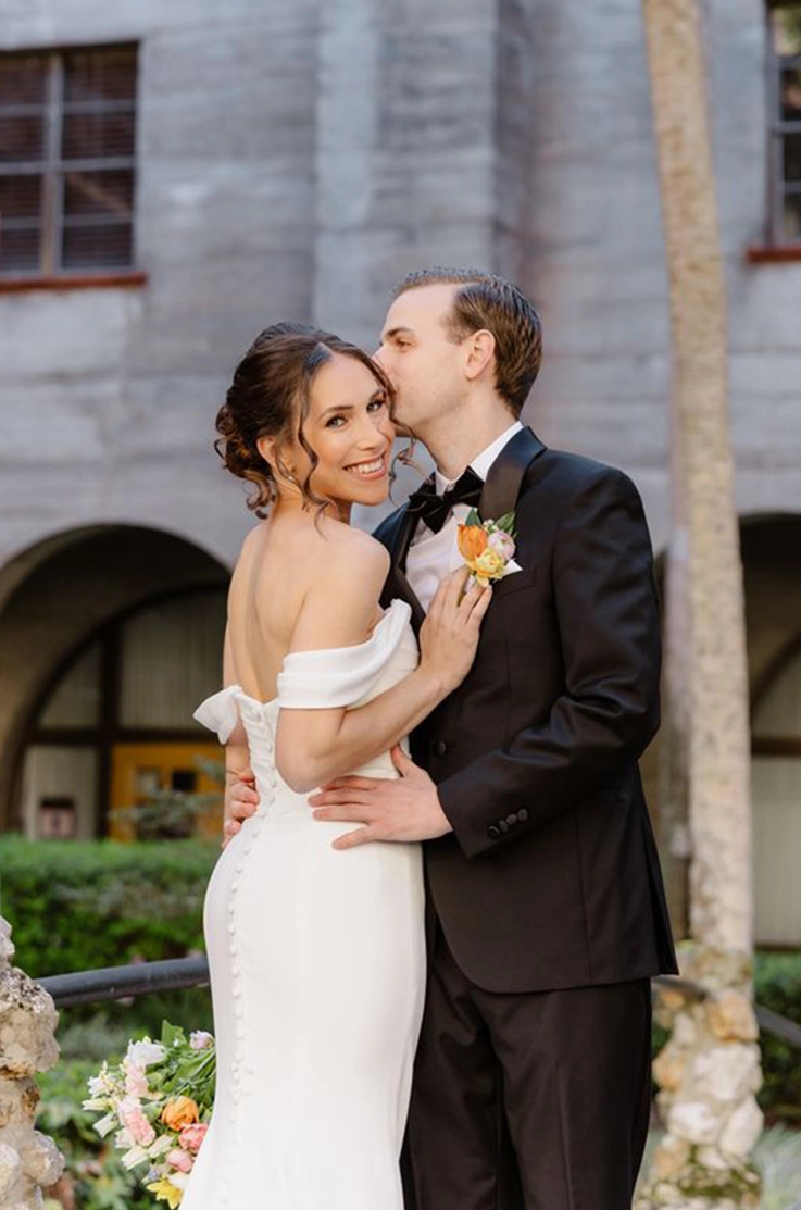  Groom holds his bride in an embrace in a crepe fitted off-shoulder gown with draped sleeves and buttons, outside the venue.