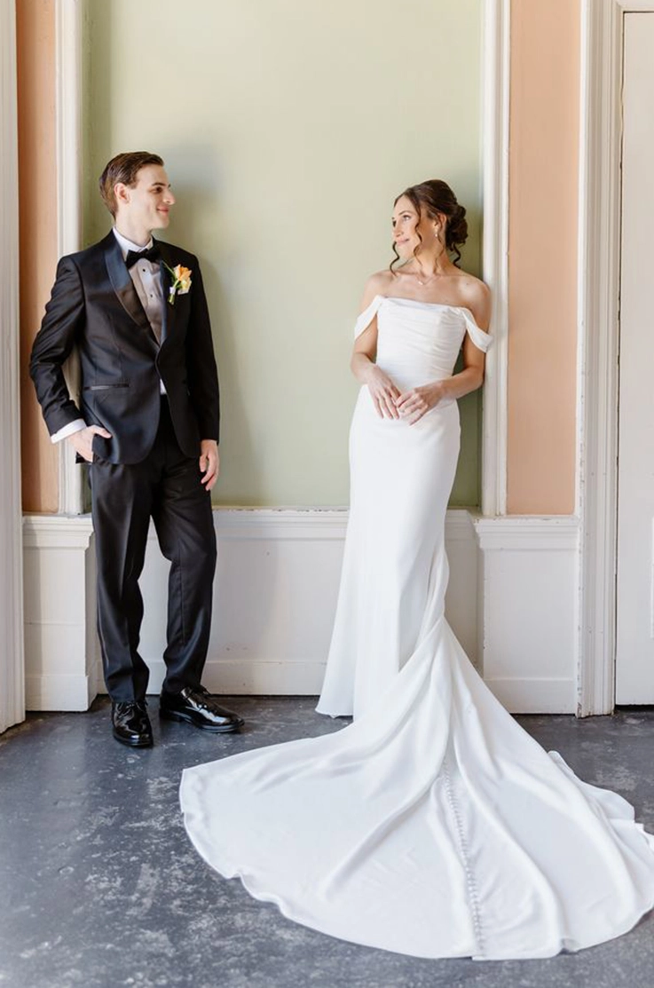Bride in a draped crepe bodice gown with sleeves and a fit and flare skirt, poses indoors and is looking at groom in a tuxedo.
