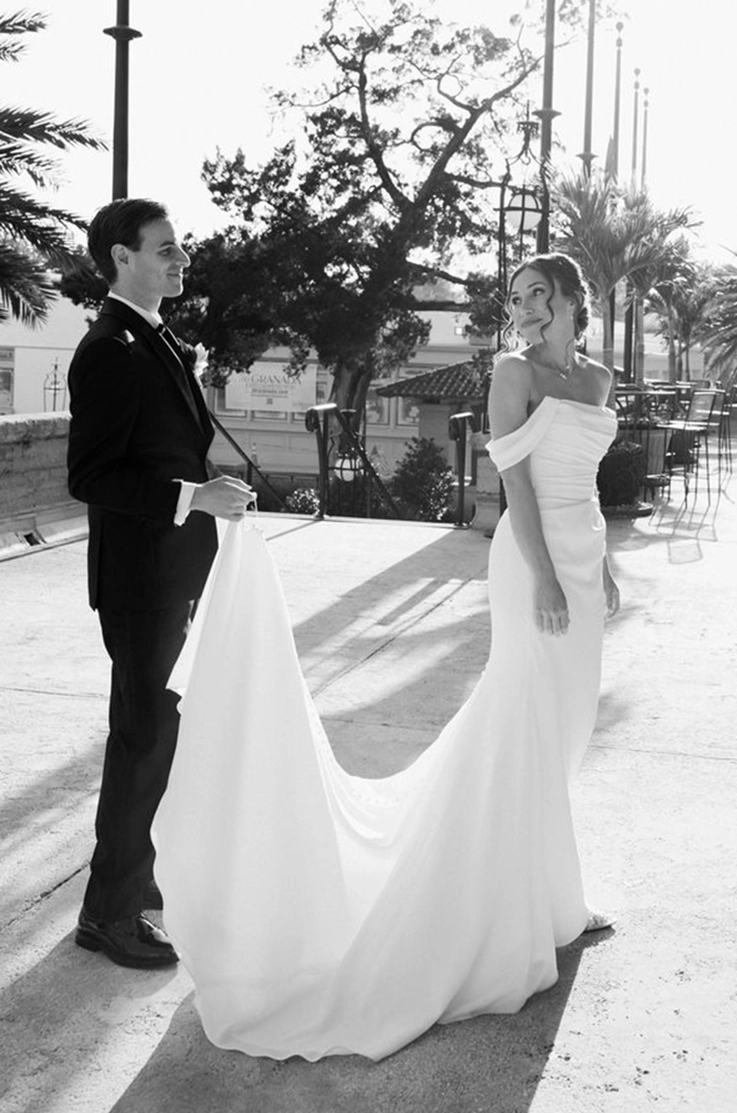Groom holds his bride's train, wearing an off shoulder crepe gown with draping and sleeves, outdoors in black and white. 