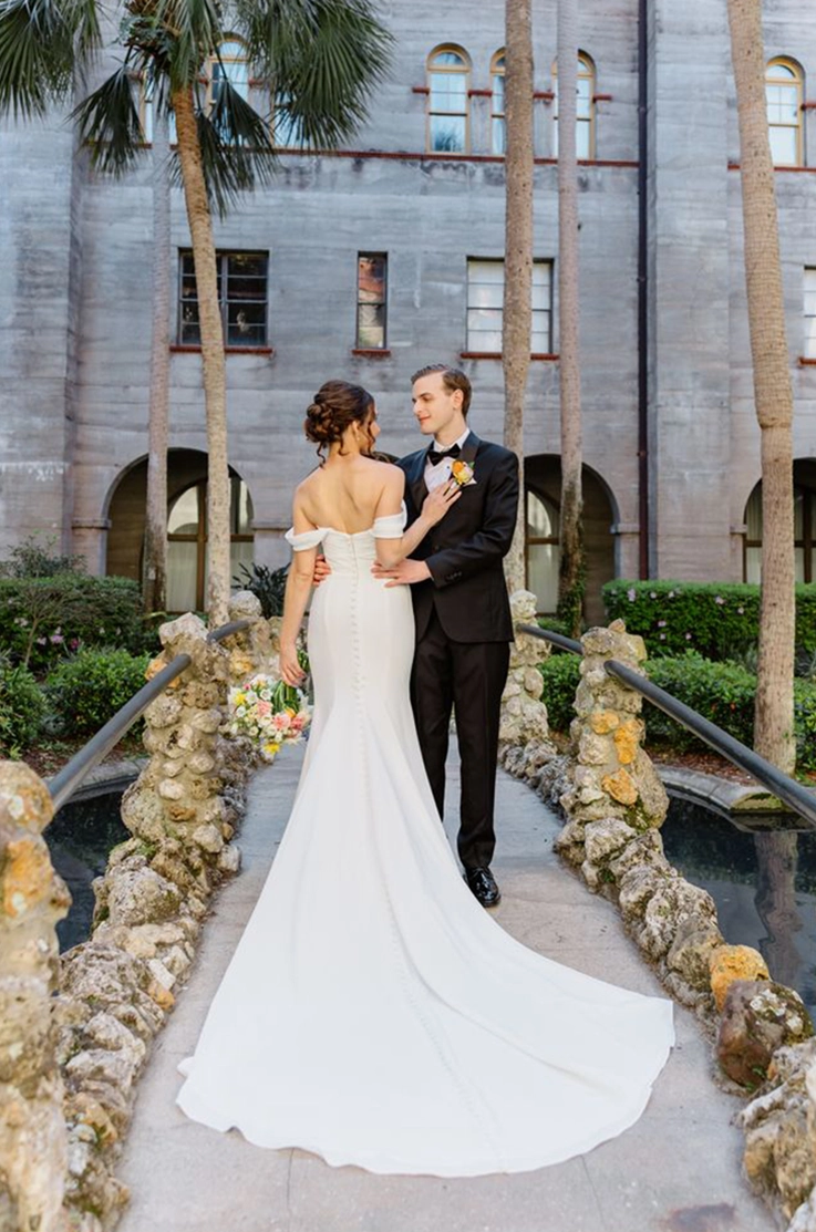 Bride in an off-shoulder fit and flare gown with buttons, stands on a bridge with groom outside the venue near palm trees. 