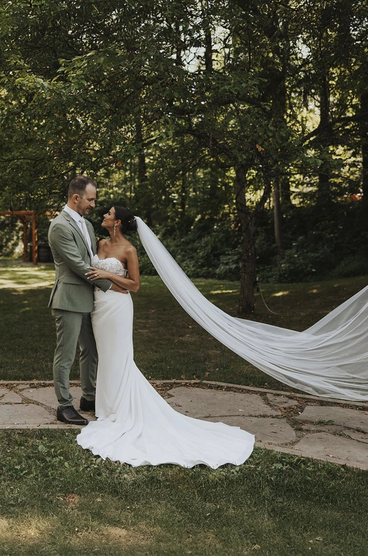 Groom embraces bride in a sweetheart neck lace gown with a crepe fit and flare skirt and a long veil, standing on a stone path.