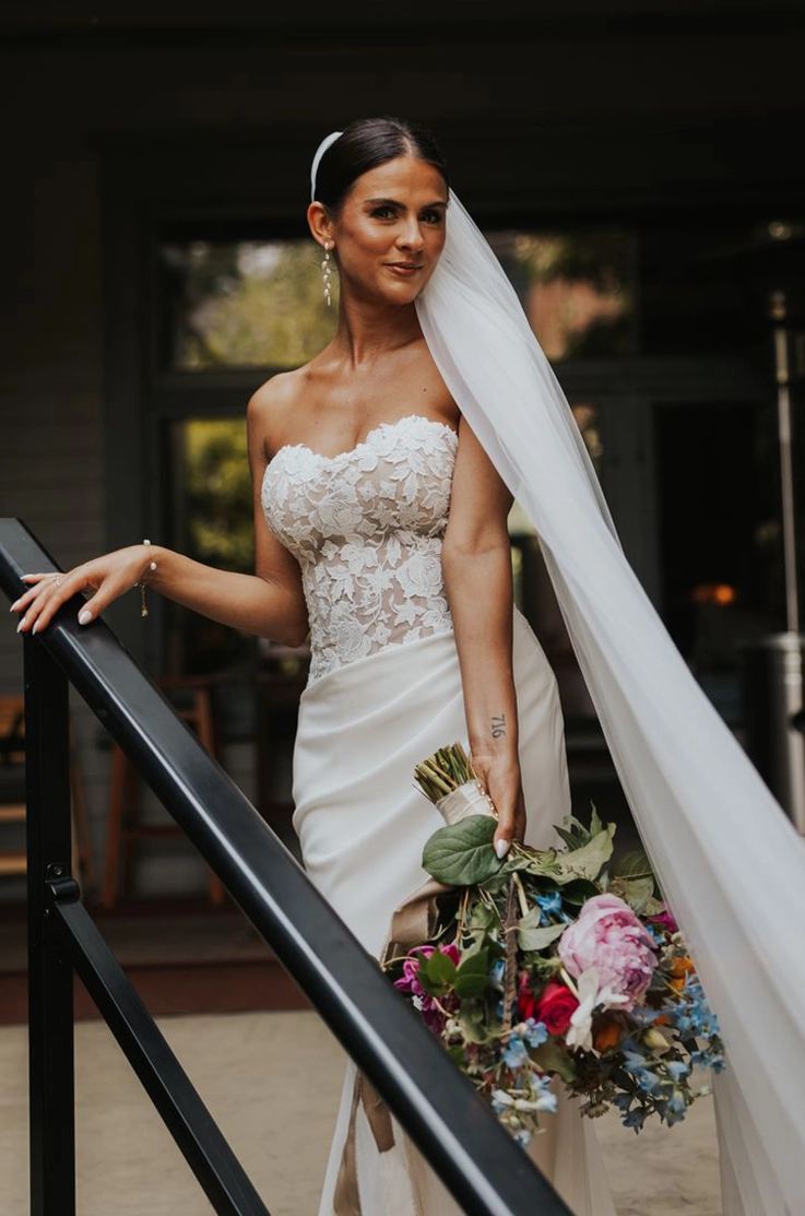 Brides smiles posing by a railing, in a strapless lace bodice wedding dress with a crepe skirt and veil, holding a bouquet.