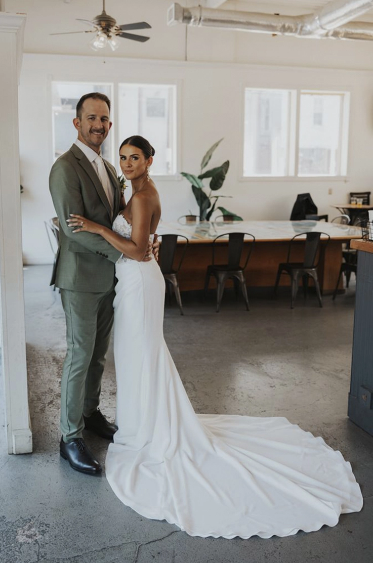 Bride in a lace bodice gown with an asymmetric crepe skirt with draping, and poses beside groom at indoor venue.