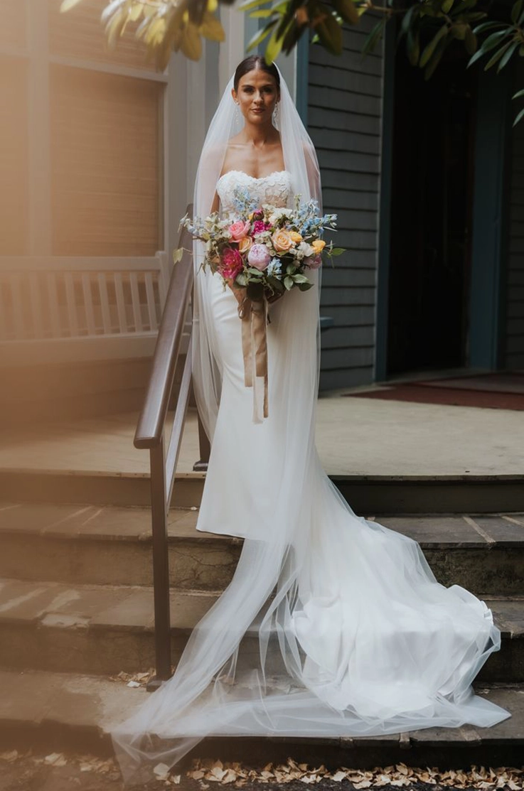 Bride standing on stone steps in a strapless lace and crepe gown with a veil and is holding a bouquet under a tree.