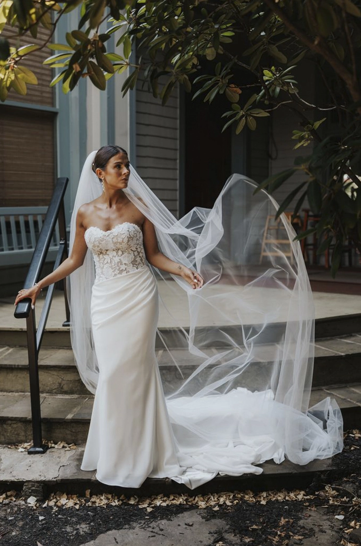 Bride in a lace sweetheart neck gown with an asymmetric crepe fitted skirt and a flowing veil, at the bottom of stone steps. 