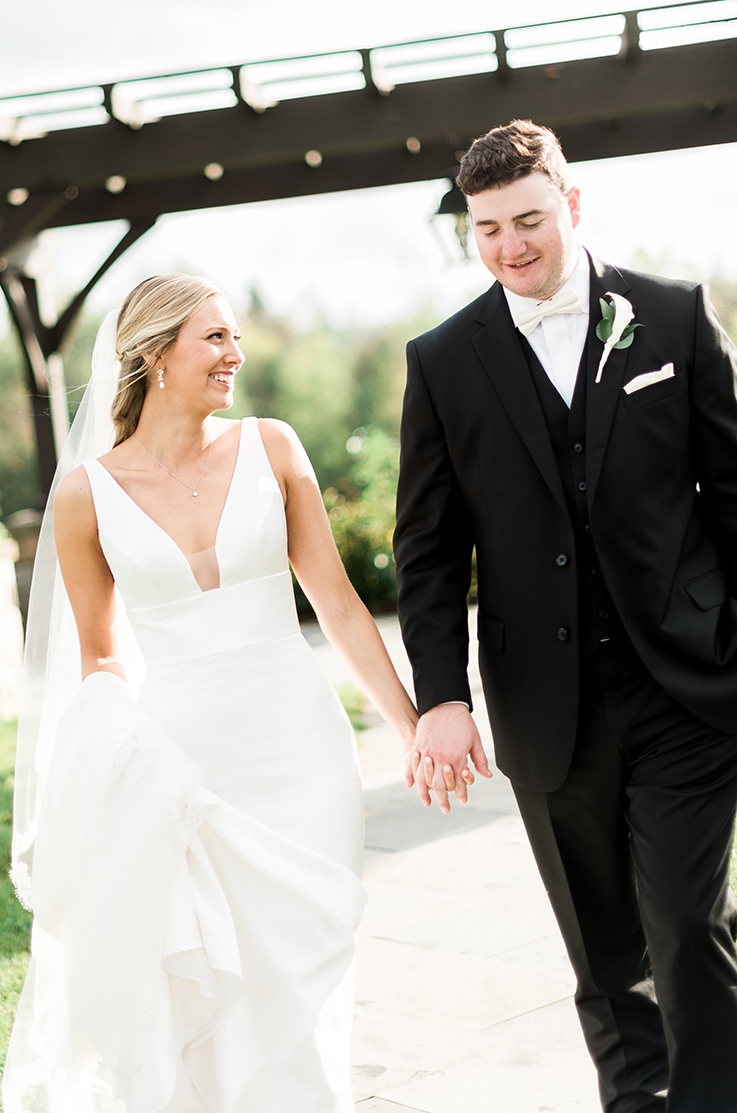 A bride in a sleeveless fitted wedding dress holds hands with a groom in a suit while they walk down a path outdoors together.