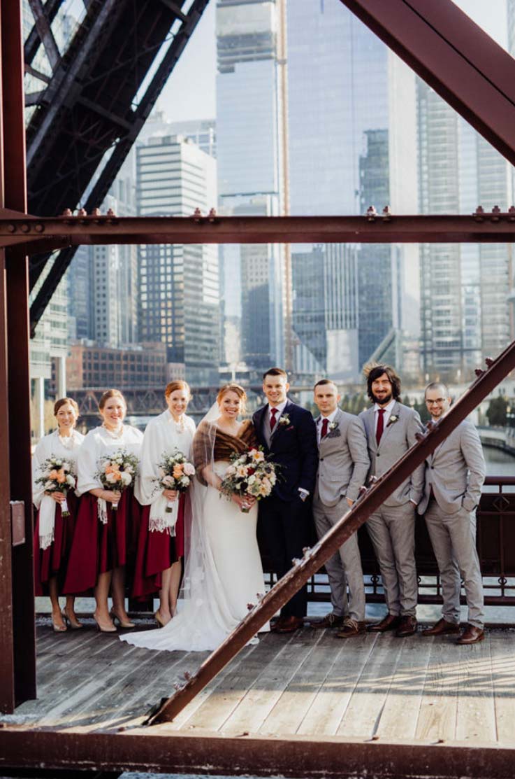 A bride in a fitted wedding dress and a fur shawl and a groom in a suit stand on a wood and rusted metal bridge with their wedding party, with a city in the background.