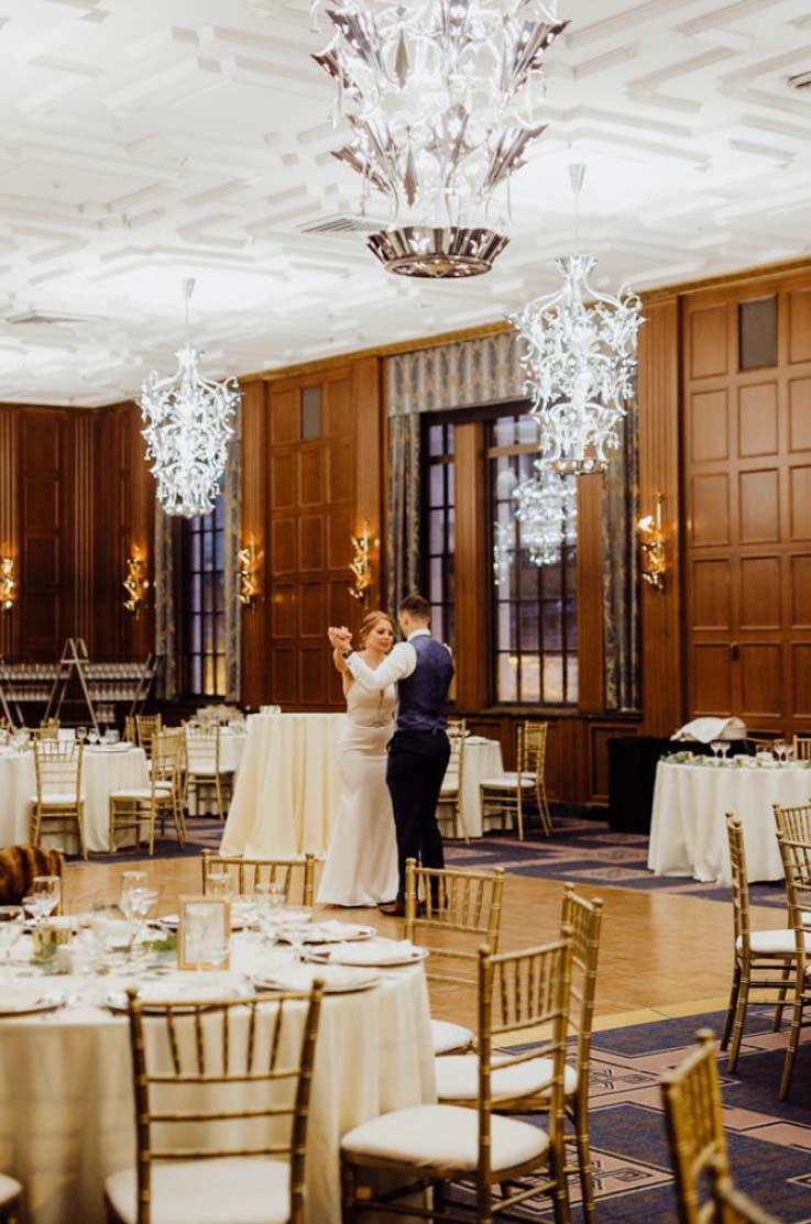 A bride in a fitted wedding dress and a groom in a suit vest dance on a dancefloor in a ballroom, surrounded by empty tables and chairs.