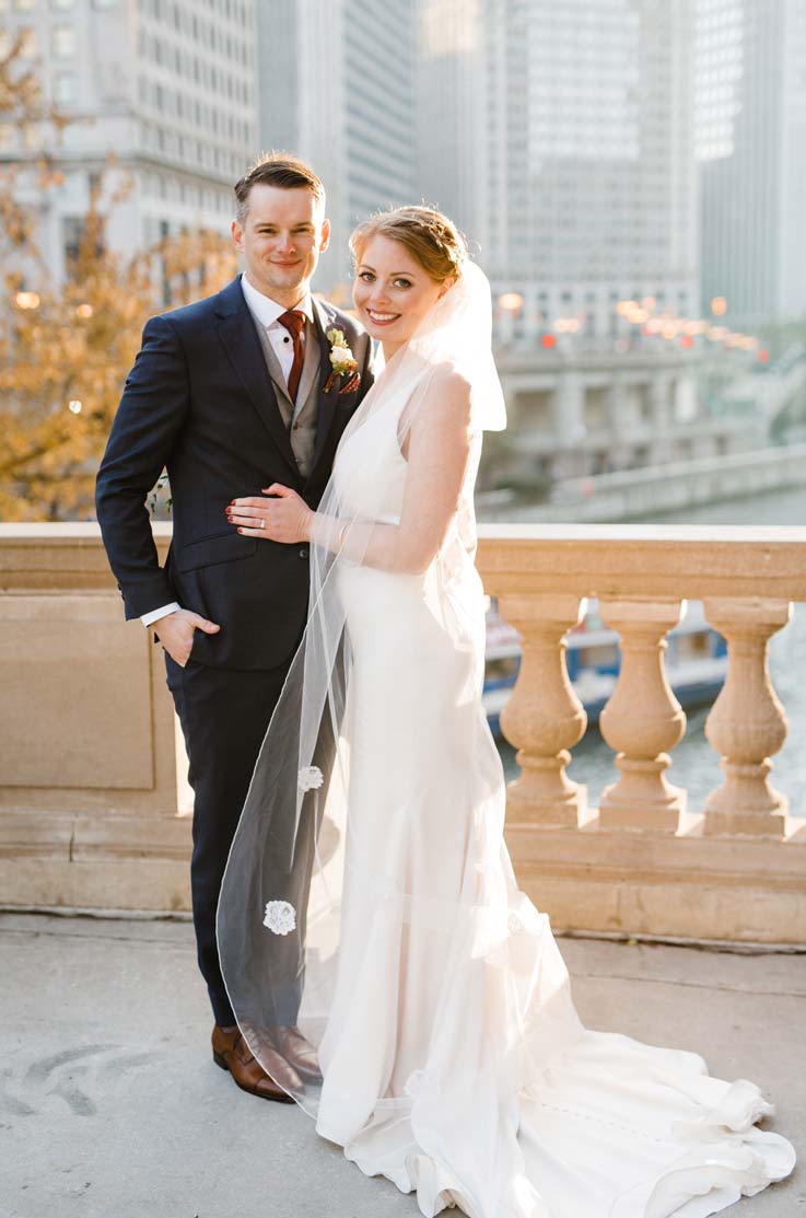A bride in a sleeveless wedding dress and a veil, and a groom in a suit stand together on a stone balcony overlooking a river and a city.