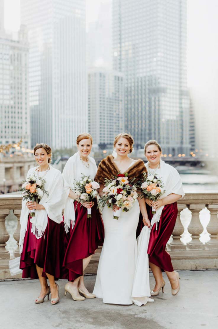 A bride in a fitted wedding dress and a fur shawl stands with three bridesmaids on a stone balcony overlooking a river and a city, while all smiling and holding bouquets.