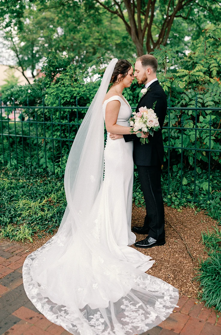 Bride in an off-shoulder fit and flare gown with buttons and a lace edge veil, faces groom and holds a blush bouquet outdoors.