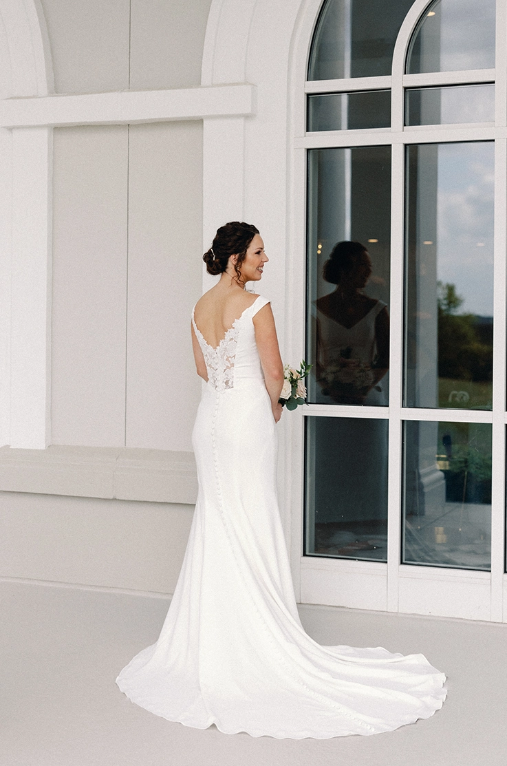 Bride poses in a crepe fit and flare wedding dress with a lace back, and is holding a bouquet outside the venue.