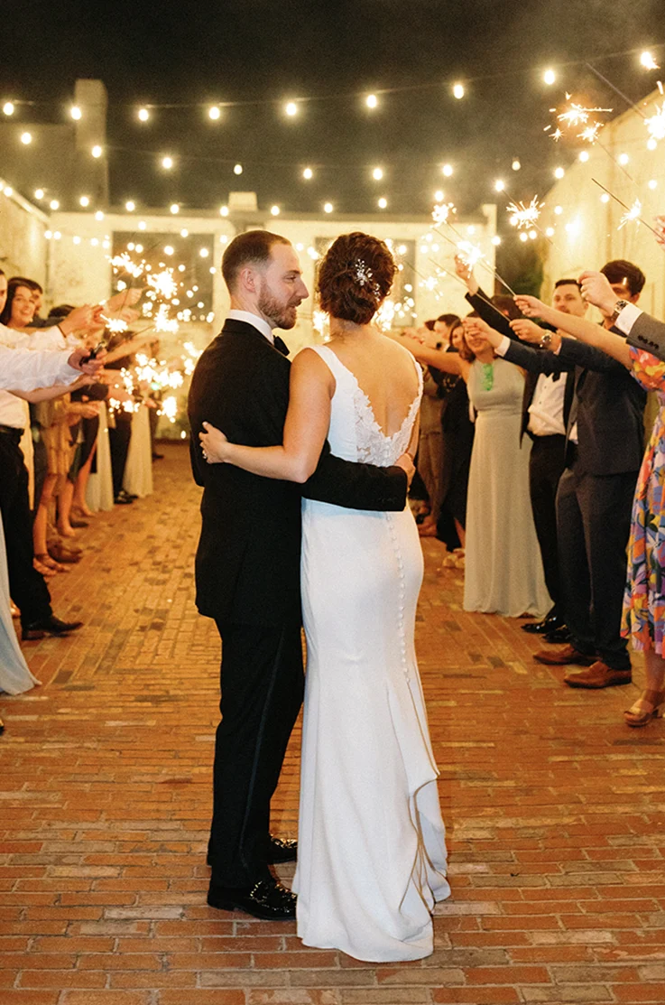 Bride in a deep-V back lace and crepe fitted gown, embraces groom near guests with sparklers under string lights. 