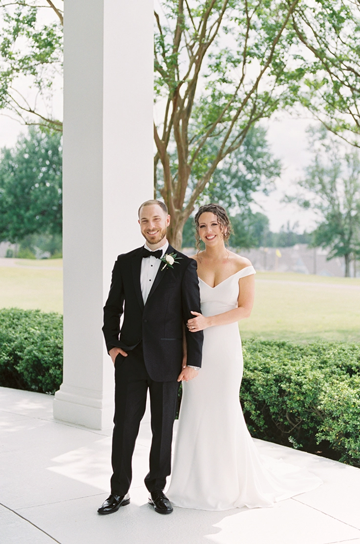 Bride in a crepe off-shoulder gown smiles next to groom in a tuxedo outside the venue on a white portico.