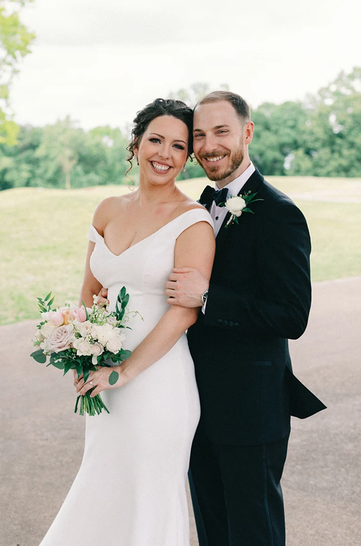 Close-up of groom embracing his bride in a crepe off-shoulder fitted gown, holding a blush bouquet on a gravel path.