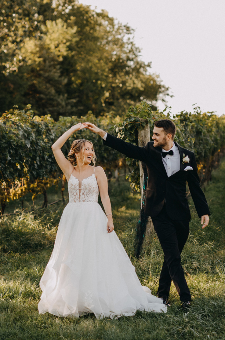 A groom in a suit smiles and spins a bride in a plunging neckline ball gown that is also smiling, while standing outdoors in front of rows of grapevines.