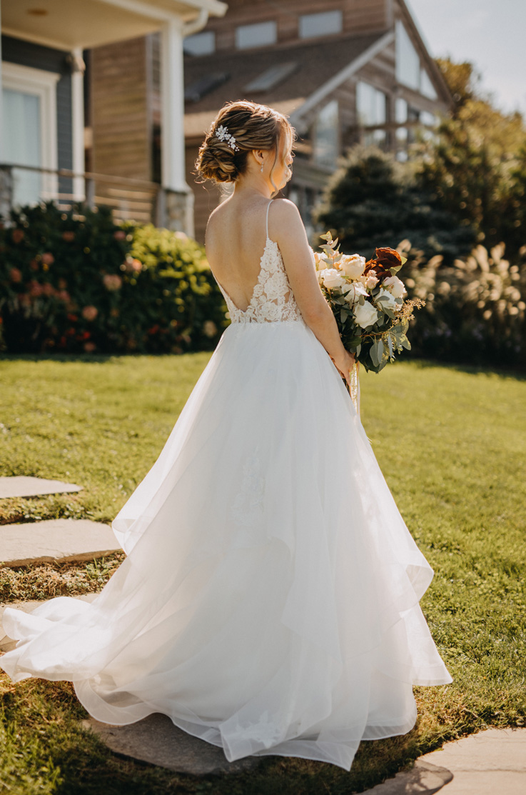 A bride in an Organza ball gown wedding dress holds a bouquet and faces away, while standing outside in the front yard of a house.