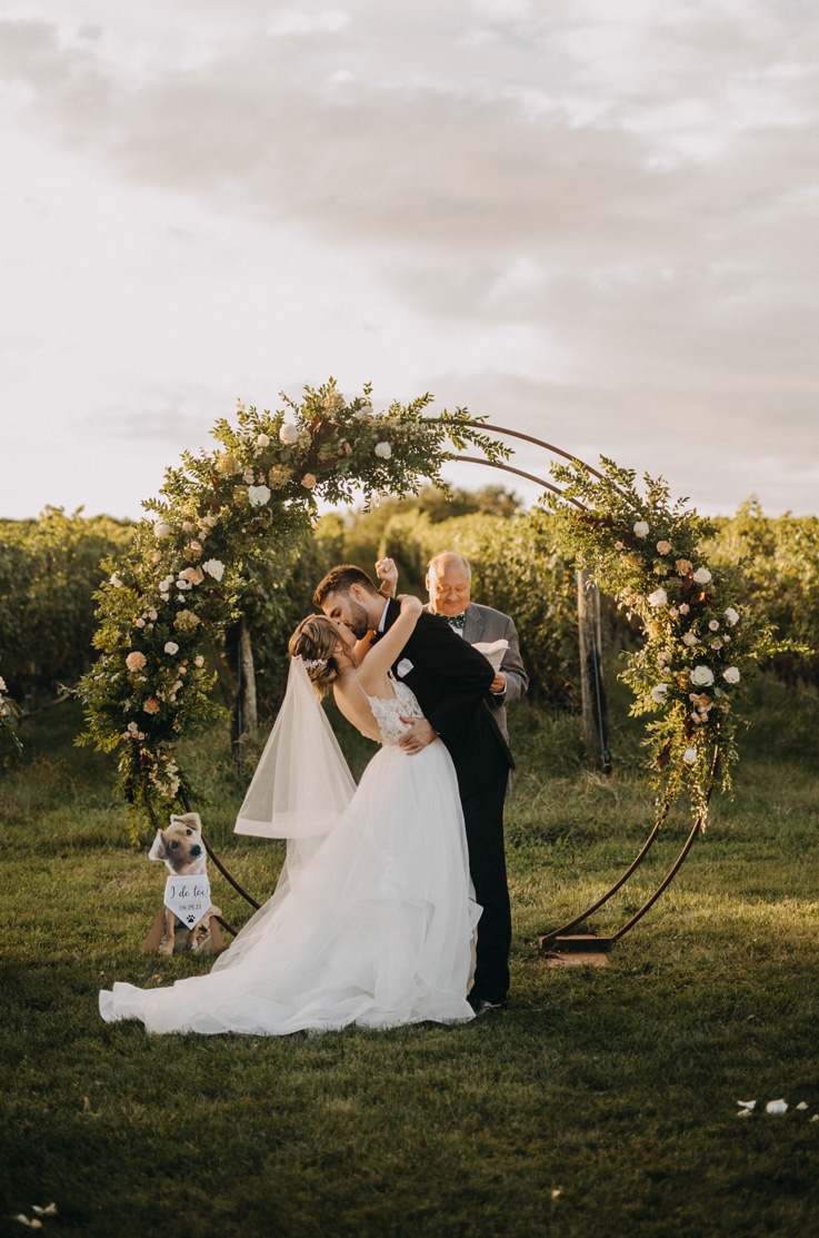 A bride in an Organza ball gown wedding dress and a groom kiss at an outdoor wedding ceremony in front of rows of grapevines an a round wedding decoration.