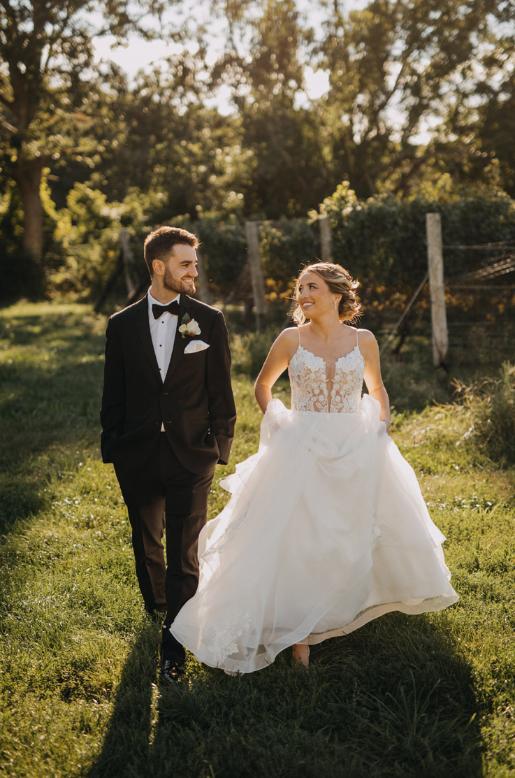 A bride in a plunging neckline ball gown and a groom in a suit walk together outside while looking at each other and smiling.