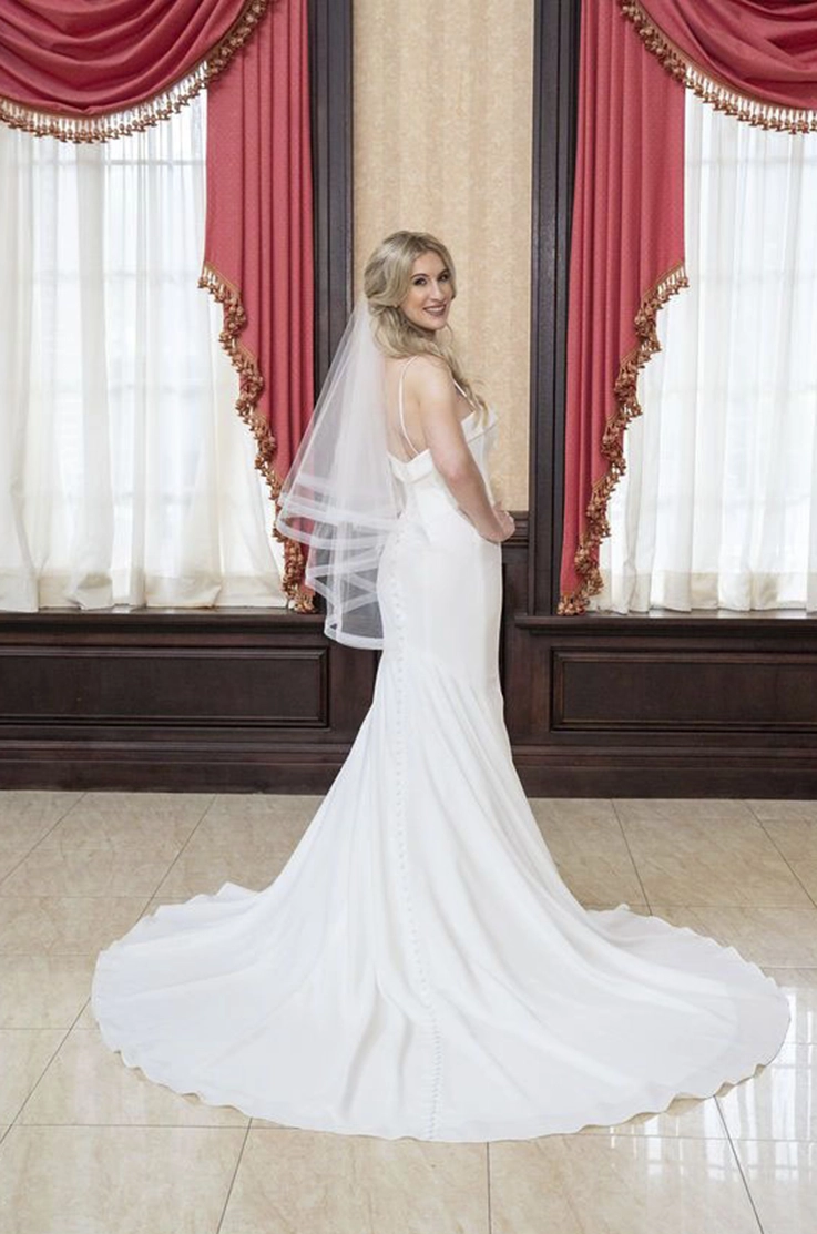 Bride in a spaghetti strap crepe gown with a fit and flare skirt and a veil, poses and looks over her shoulder in a ballroom.