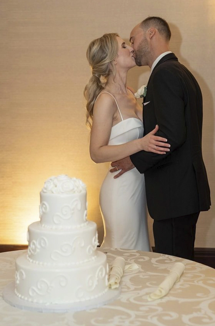 Groom kisses his bride in a low back crepe fitted gown with spaghetti straps, by a three-tier cake at indoor reception.