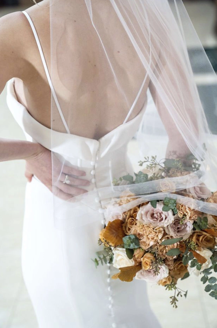 Close-up detail of bride in a low back buttoned crepe gown with thin straps and a veil, is holding flowers by her back.