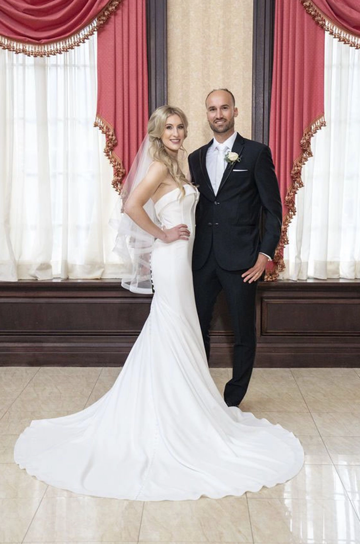 Bride in a fit and flare crepe wedding dress with a veil and buttons down the back, poses with groom in an elegant ballroom.