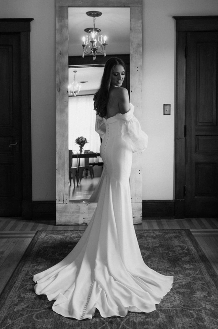 Bride glances over her shoulder, in front of a mirror in a crepe fitted and buttoned gown with sleeves, in black and white.