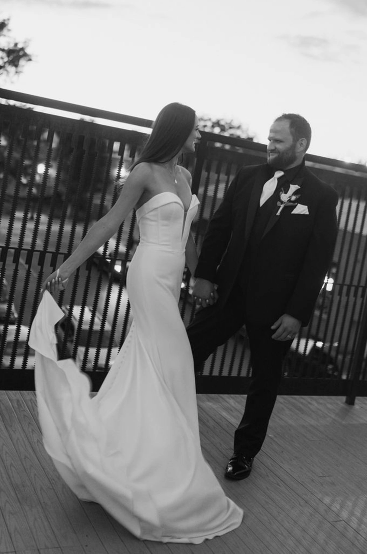Bride picks up her train in a strapless fit and flare gown with a sweetheart neck, beside her groom, in black and white.