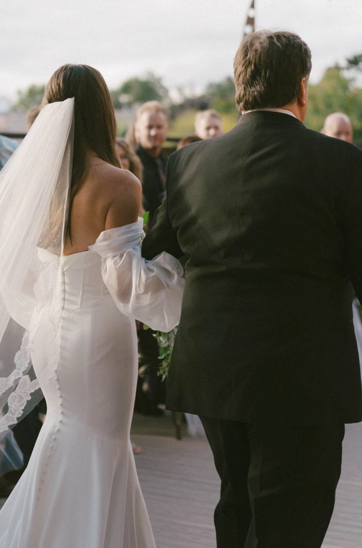 Back of bride in a fit and flare crepe gown with puff sleeves and a lace-edged veil, walking down the aisle in outdoor ceremony.
