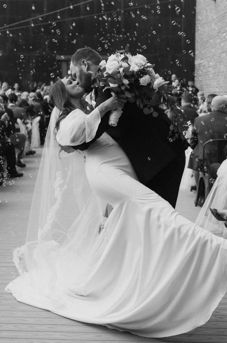 Groom dips his bride in a crepe fitted gown with a lace-edged veil and they kiss in the aisle, with bubbles floating.