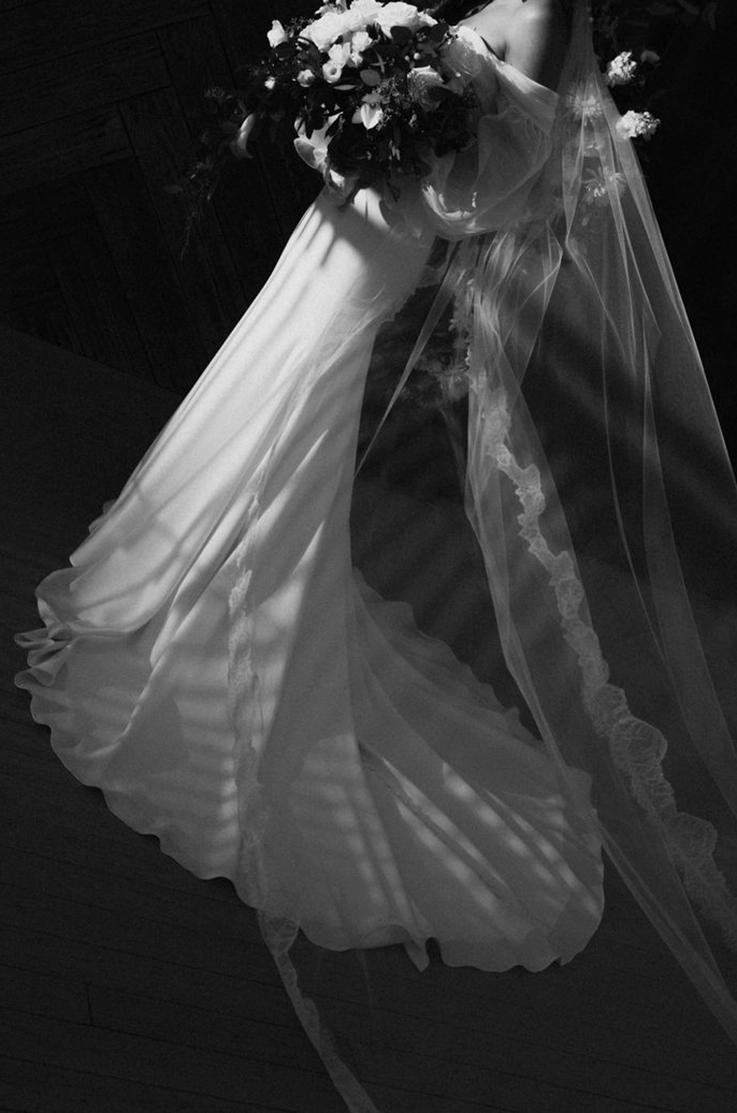 Bride poses in a crepe fit and flare wedding dress with a lace-trim veil and is holding a bouquet, shown in black and white.