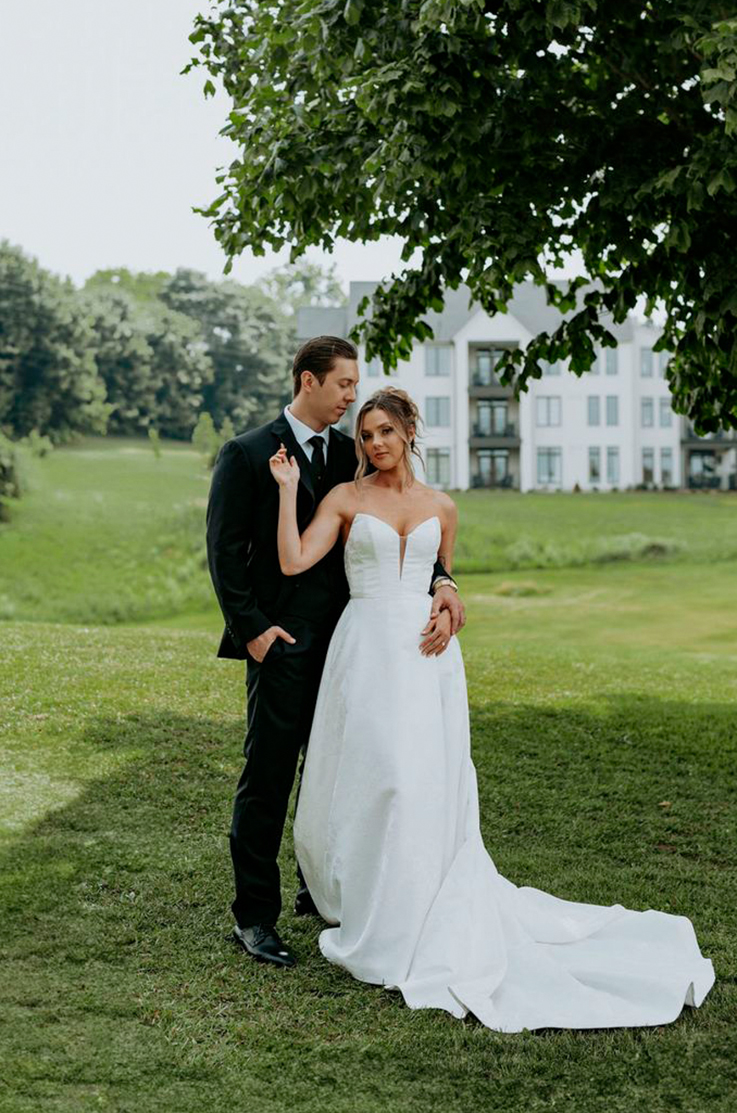Bride in a jacquard print A-line wedding dress with a plunging neckline, poses alongside her groom on a sunny lawn.