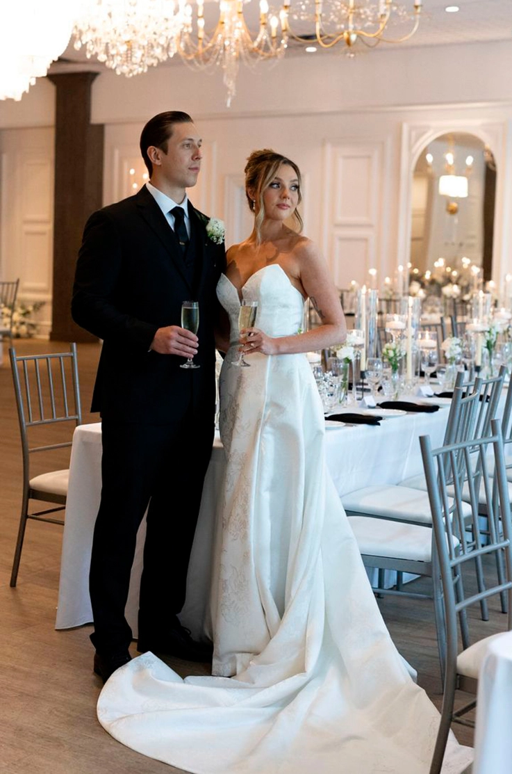 Bride in a plunging neck jacquard print gown poses next to groom, in indoor reception under lit chandeliers.