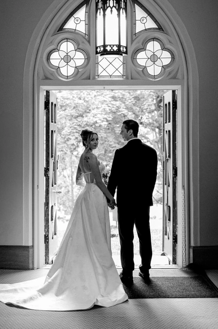 Bride in a strapless jacquard seamed bodice gown, holds hands with groom in an arched open doorway.