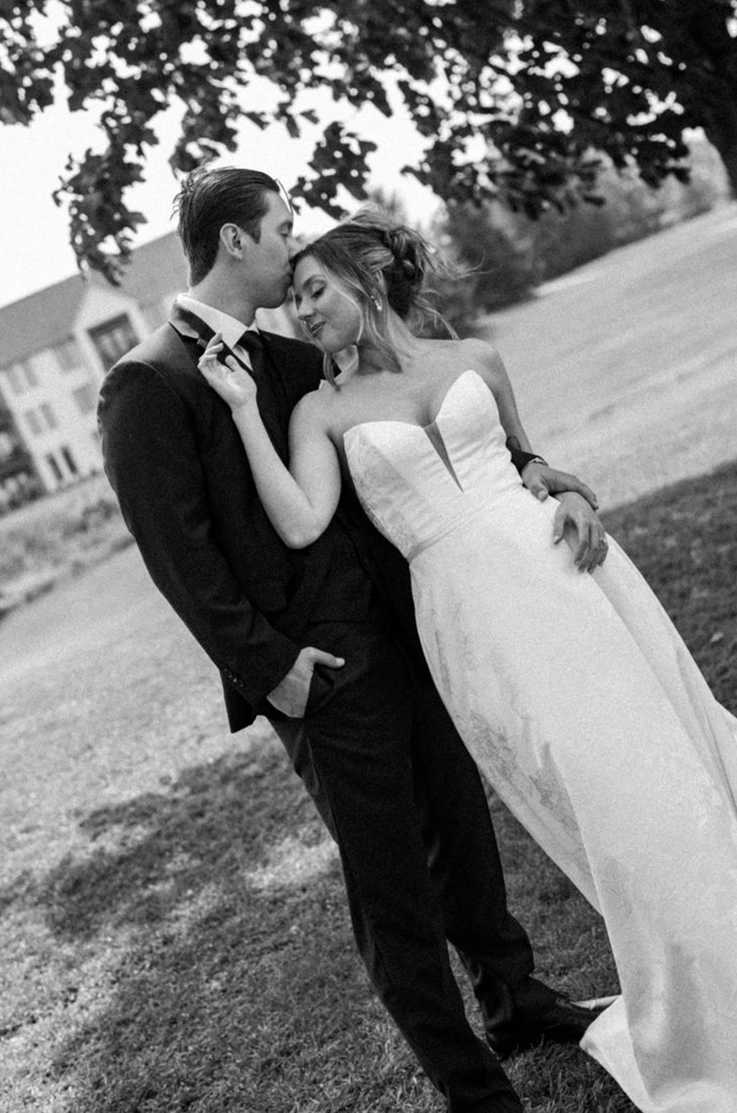 Groom embraces bride in a strapless plunging neck gown with a jacquard skirt, under a tree on a lawn in black and white.