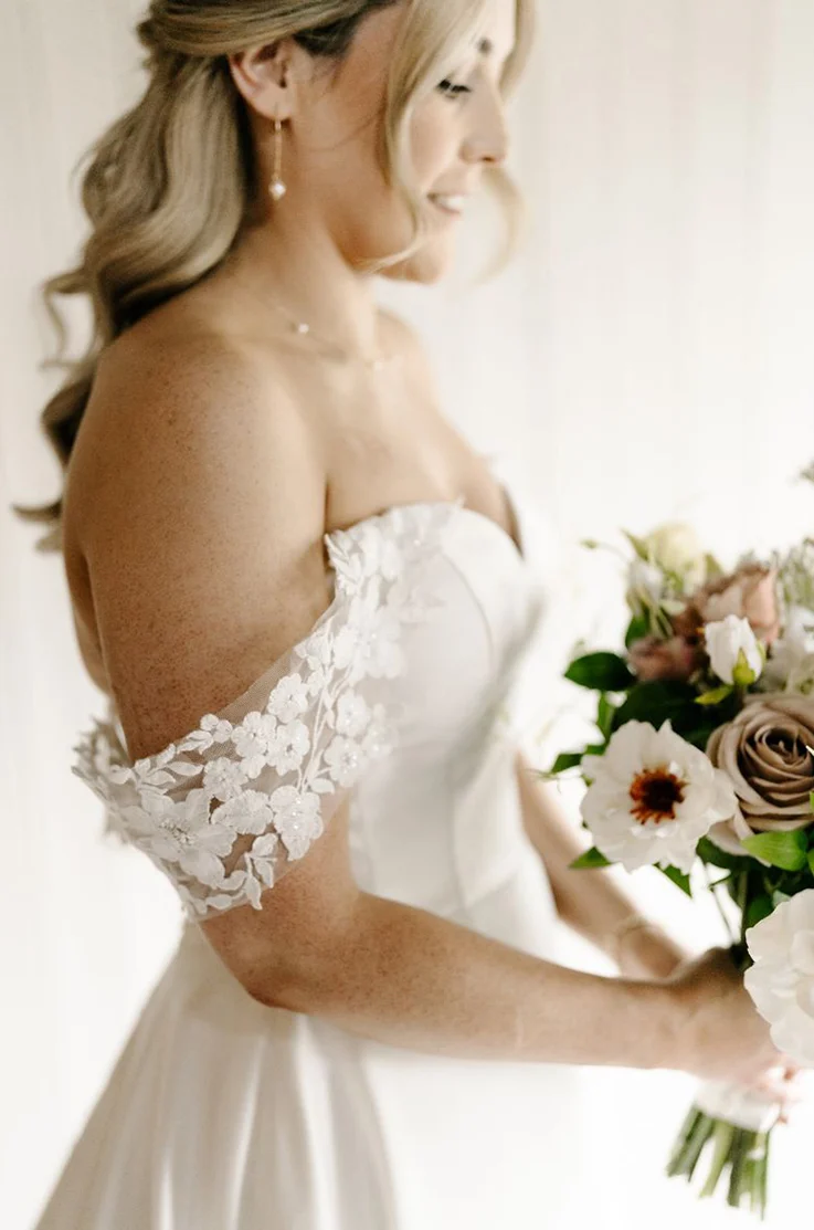 Close-up of bride in an off-shoulder floral lace and mikado wedding dress, is holding a bouquet indoors in soft lighting. 