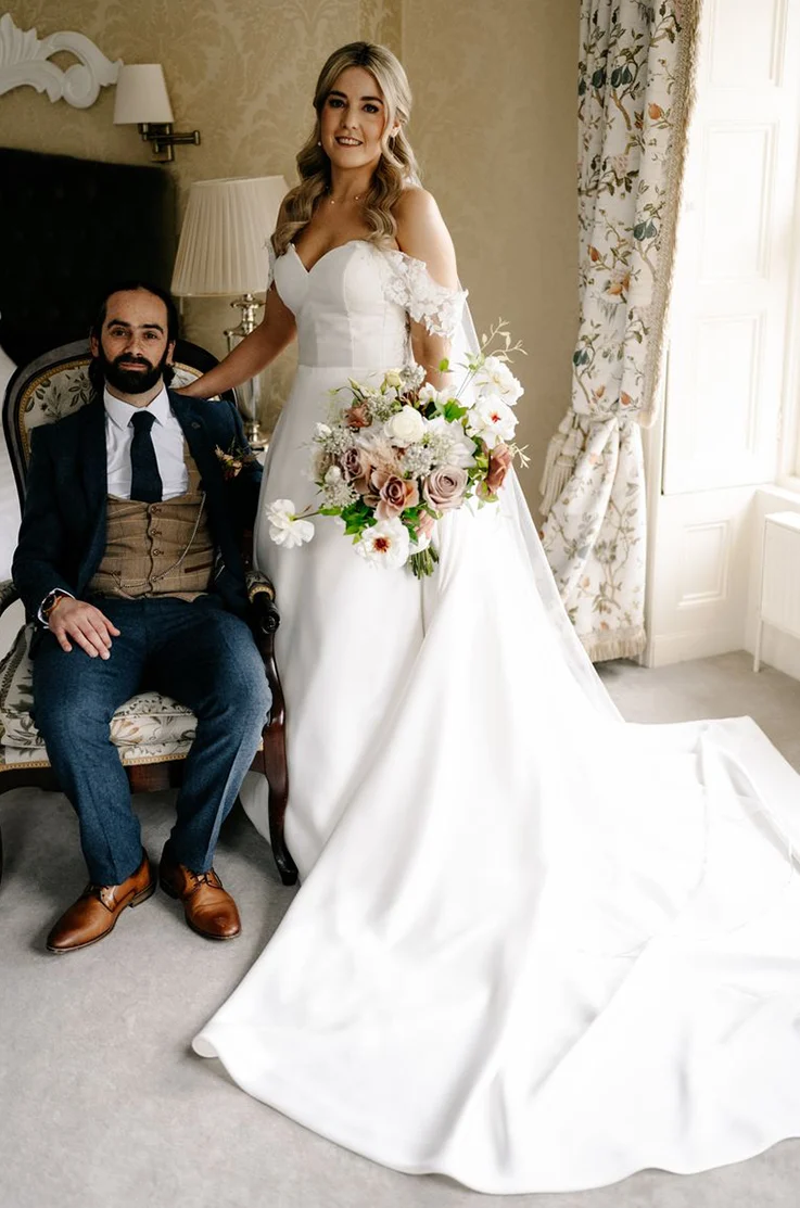 Bride in a strapless mikado gown with floral lace sleeves, poses beside groom, holding a large bouquet in an elegant manor.