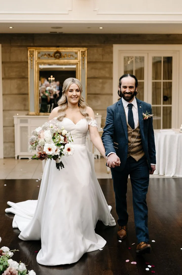 Bride in a sweetheart neck mikado gown with lace sleeves, is holding a bouquet and walks hand in hand with groom in the aisle. 