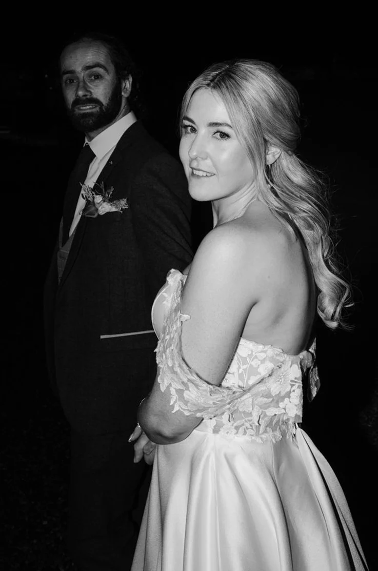 Bride in an off-shoulder floral lace bodice gown with a full mikado skirt, standing next to her groom, in black and white. 