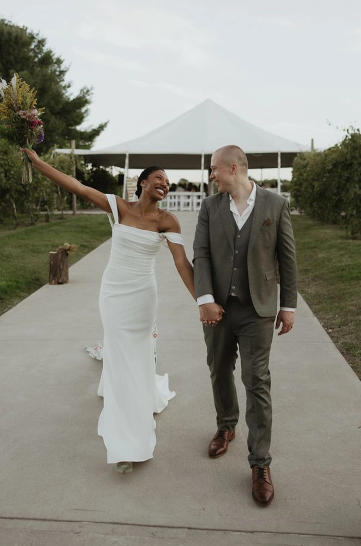 Bride smiles in an off-shoulder draped crepe wedding dress, walking hand in hand with groom at outdoor wedding by a tent.