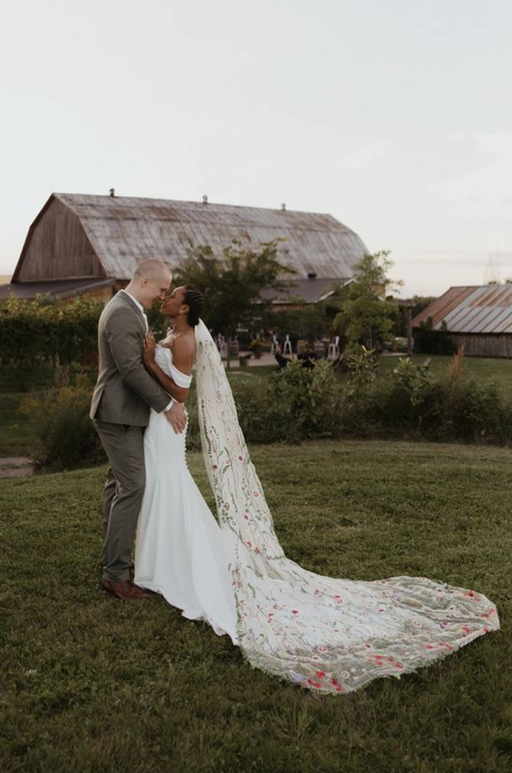 Groom embraces bride in a crepe fit and flare gown with a buttoned back, draped sleeves and an embroidered veil by a barn. 