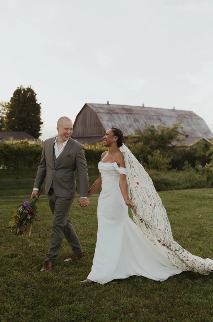 Bride in a draped crepe bodice gown with sleeves and a flowing veil, walks hand in hand with groom at an outdoor barn.