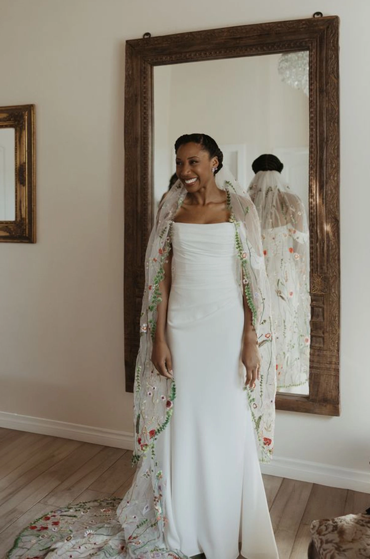 Bride in a scoop neck crepe fitted gown with a draped bodice and embroidered veil, smiling by an ornate mirror indoors.
