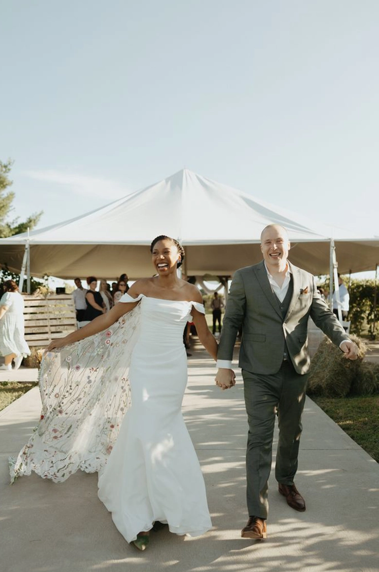 Bride in a draped crepe bodice gown with sleeves, and a veil, holds hands celebrating with groom by an outdoor tent.