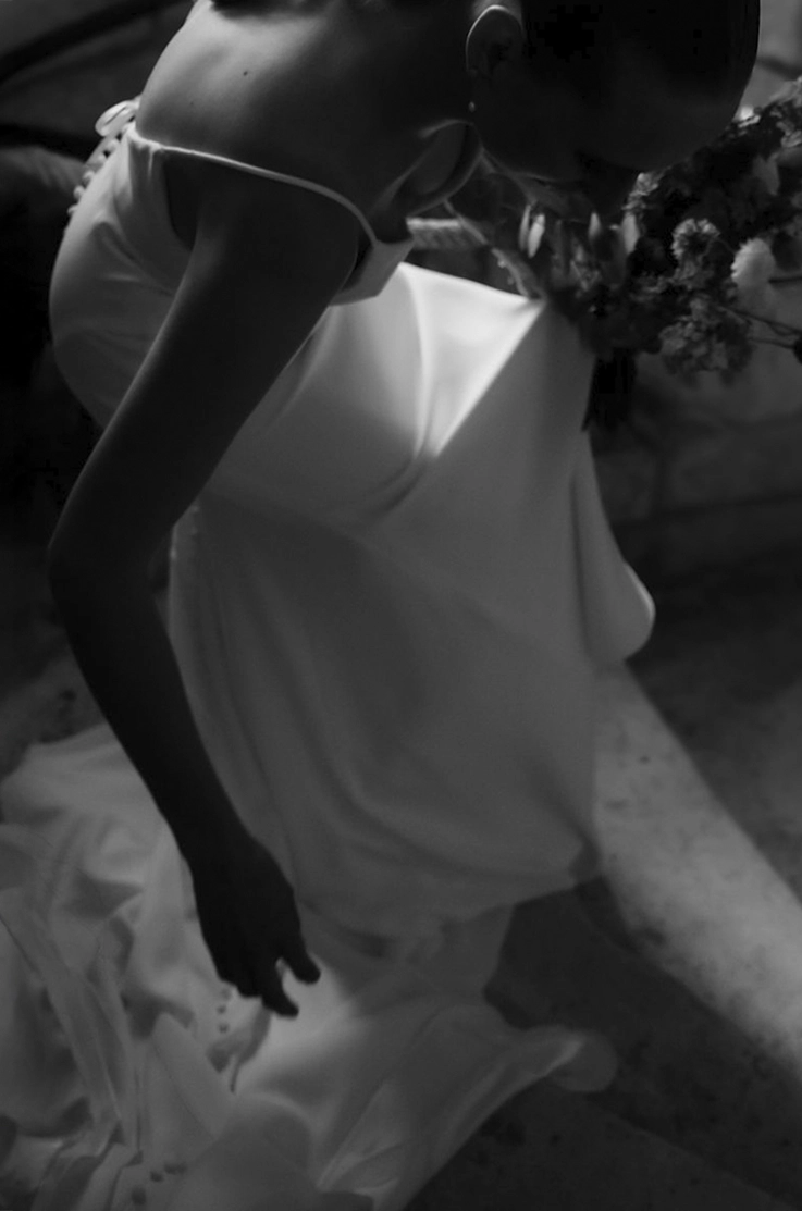 Bride in a spaghetti strap crepe wedding dress with a buttoned back and sash, holding a bouquet and shown in black and white. 
