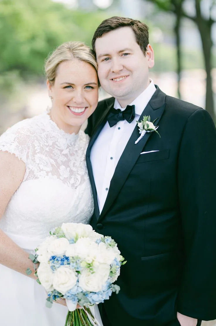 Close-up of bride smiling in a lace bodice wedding dress with a jewel neck and holds a bouquet, beside her groom outdoors.