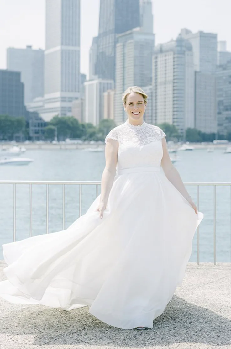Bride twirls in a lace and organdy A-line gown with a jewel neckline, smiling at a waterfront in front of a city skyline.