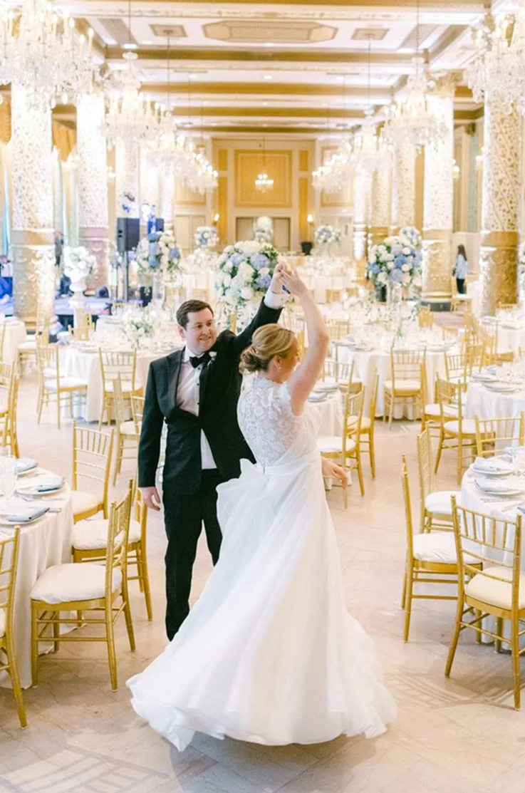 Groom twirls his bride in a lace bodice gown with an organdy ribbon and skirt, in a ballroom with gold chandeliers.