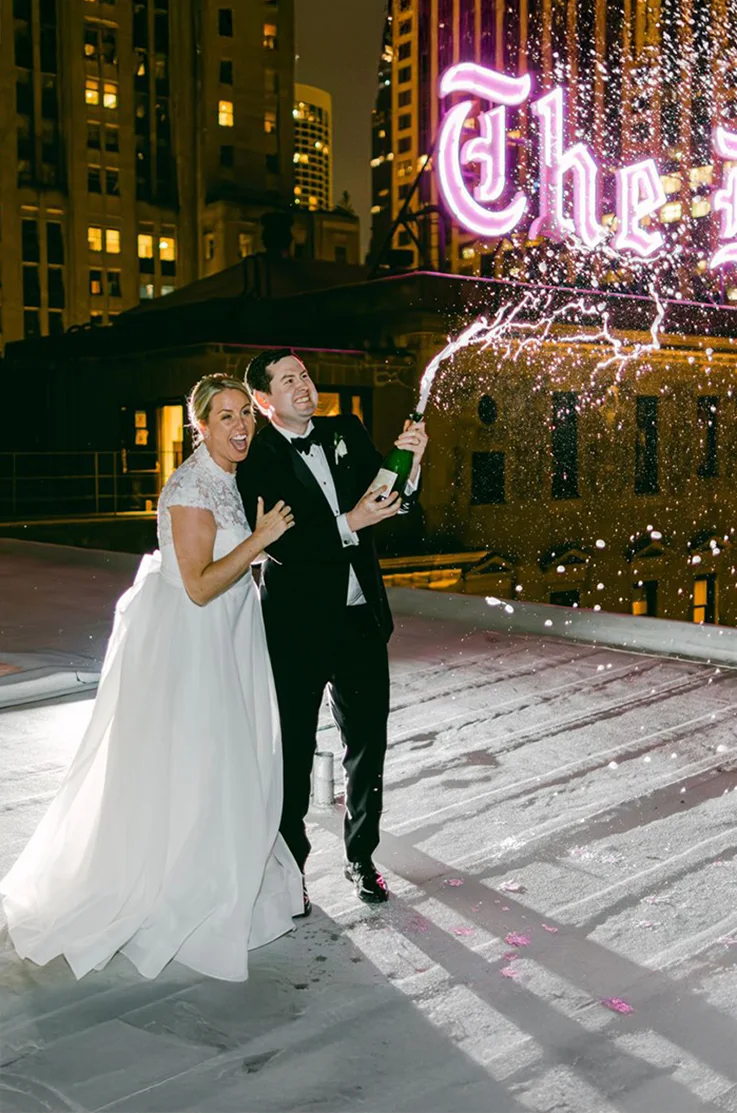 Bride in a lace and organdy A-line gown celebrates alongside her groom, popping champagne on a city rooftop at night.